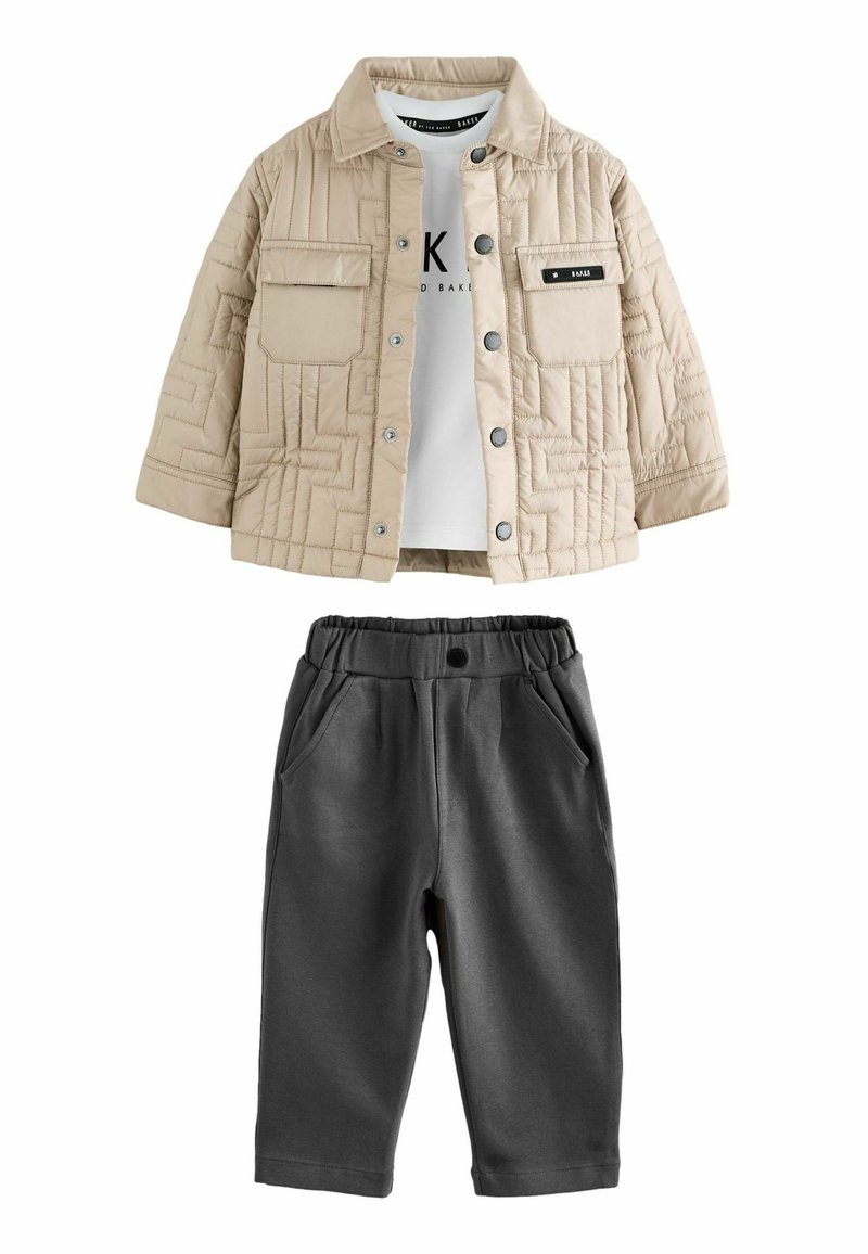 Beige quiltet jakke med trykknapper over hvid T-shirt med logo, parret med mørkegrå elastiske bukser med forlommer.