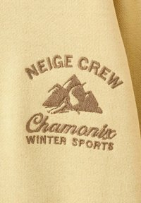 Krémově zbarvená látka s hnědou vyšívanou nášivkou s horami a textem: "NEIGE CREW Chamonix ZIMNÍ SPORTY."