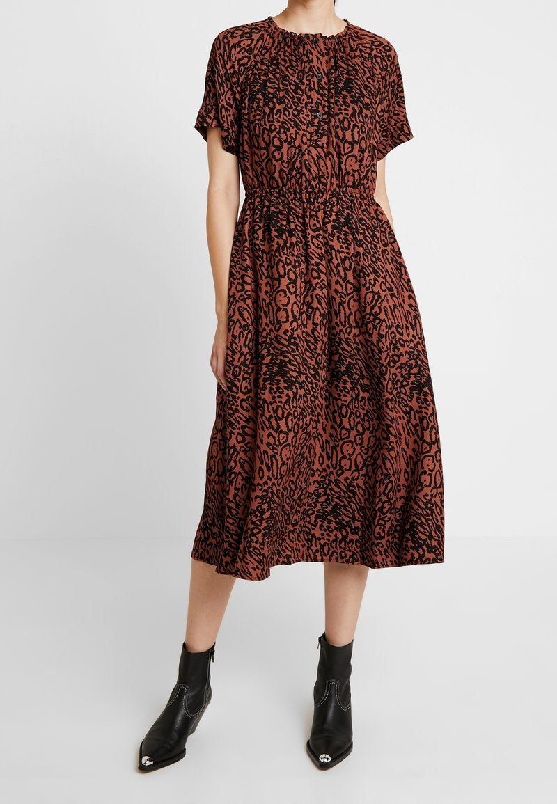 Braunes Tiermusterkleid mit kurzen Ärmeln, gerafftem Ausschnitt, elastischem Bund und fließendem Midilängenrock, kombiniert mit schwarzen Knöchelstiefeln.