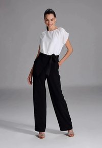 Naine seisab heledates lühikestes varrukates bluusides ja kõrge vöökohtadega mustades laiades pükstes, millel on vöö, naeratades ühtlasel hallil taustal.