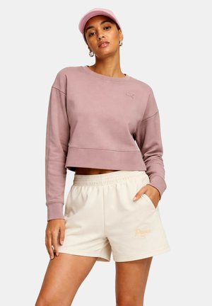 Kvinde iført lilla cropped sweatshirt, lyserød kasket og cremefarvede Puma-shorts med elastisk talje, stående med den ene hånd i lommen.