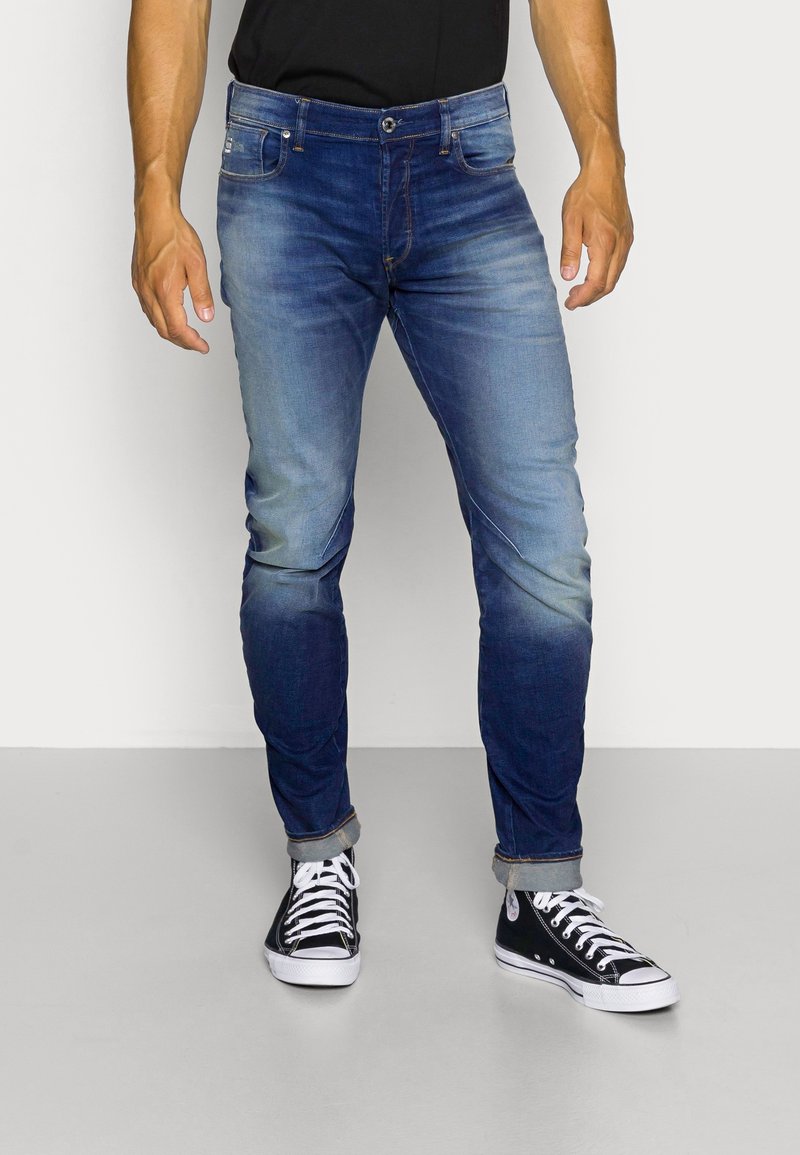 Blå jeans med en urtvättad design, slim fit och sydda detaljer, tillsammans med svarta högklackade sneakers, uppvisade mot en neutral bakgrund.