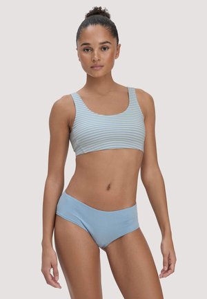 Frau trägt hellblau gestreiftes Crop-Top und einfarbige blaue hochgeschnittene Unterwäsche, steht vor einem schlichten hellen Hintergrund.