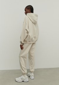 Beige, oversized kapucnis pulóver, kényelmes textúrával, hozzá illő, taperd fazonú melegítőnadrággal és fehér sportcipővel, hátulról nézve, semleges háttér előtt.