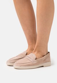 Mocassini beige in suede con suole in gomma basse, indossati a piedi nudi, che mostrano dettagliati punti di cucitura e una fascia per monete su sfondo bianco.