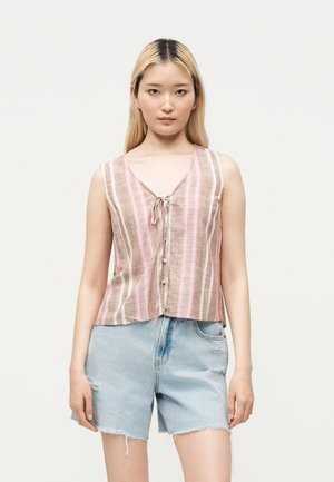 Jeune femme aux cheveux blonds et lisses portant un débardeur rayé rose et beige et un short en jean bleu clair.