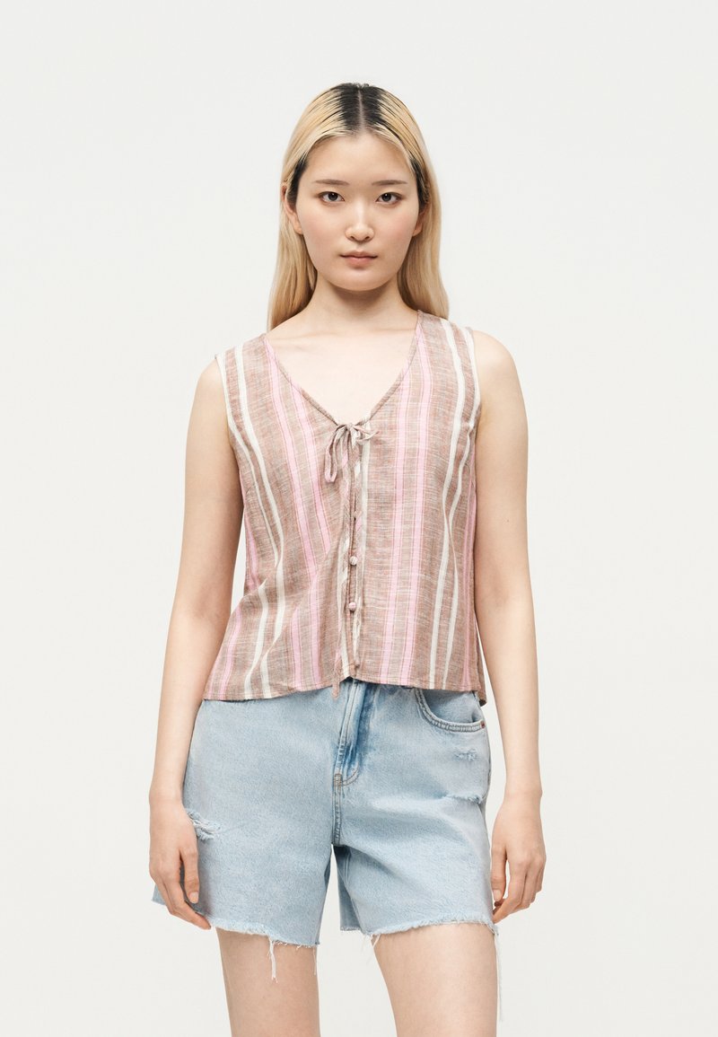 Jeune femme aux cheveux blonds et lisses portant un débardeur rayé rose et beige et un short en jean bleu clair.