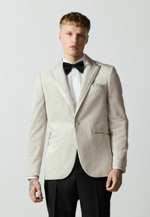 Junger Mann trägt eine weiße Smokingjacke, schwarze Fliege, weißes Hemd und schwarze Hose, steht vor einem einfarbigen grauen Hintergrund.