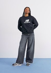 Zwarte sweatshirts met witte "New Balance" logo, gecombineerd met losse, donkere spijkerbroeken en zwart-witte sneakers. Gladde stof, relaxte pasvorm.