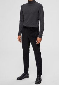 Svart polotröja i en figurnära design, kombinerad med svarta slim-fit byxor. Båda plaggen är tillverkade av slät, mjuk tyg. Skorna är i svart läder.