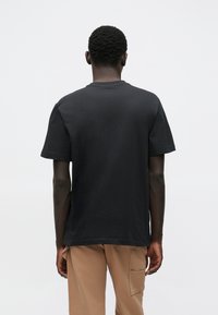 Black cotton t-shirt with a round neckline and short sleeves, displayed from the back, paired with beige cargo pants.