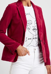 Henkilö, joka on pukeutunut punaiseen samettitakkiin, valkoisiin housuihin ja valkoiseen t-paitaan, jossa on jatkuvalla viivalla piirretty kasvokuvio ja teksti "Olen omaani."