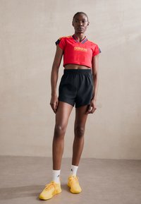 Red cropped sports jersey with yellow logo, paired with black athletic shorts. Yellow sneakers with white socks featuring logo. Neutral background.