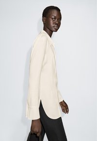 Lysebeige skjorte med knapper, lange ærmer og sideslids, lavet af glat stof. Båret med sorte bukser og en tekstureret håndtaske.