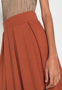 Lähikuva henkilöstä, jolla on päällään tekstuurinen hihaton paita, joka on työntynyt korkeavyötäröisten, laskostettujen ruosteenväristen hameen sisään, käsi rentona sivulla.