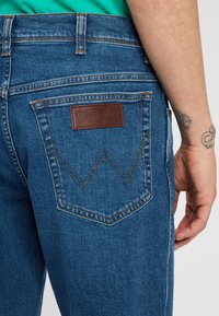 Jeans in denim blu con una patta in pelle Wrangler e cuciture distintive sulla tasca posteriore. Presentano un taglio dritto e una tonalità di lavaggio medio.