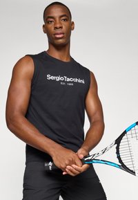 Svart ermelig t-skjorte med hvit tekst "Sergio Tacchini EST. 1966". Modellen holder en tennisracket med blå aksent og har på seg sorte shorts.