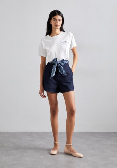 Weiße Baumwoll-T-Shirt mit gesticktem Text, marineblaue Denim-Shorts mit einem gemusterten Stoffband, und nudefarbene flache Schuhe mit Knöchelriemen.