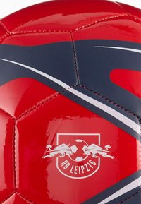 Balón de fútbol rojo con una superficie lisa y brillante, con acentos curvos en azul oscuro y blanco, y el logo de RB Leipzig impreso en blanco.