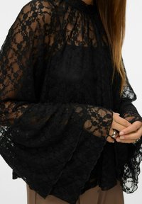 Svart bluse i blonder med høy hals, flagrende klokkeformede ermer og et lagdelt design med blomsterpatroner og gjennomskinnelig tekstur.