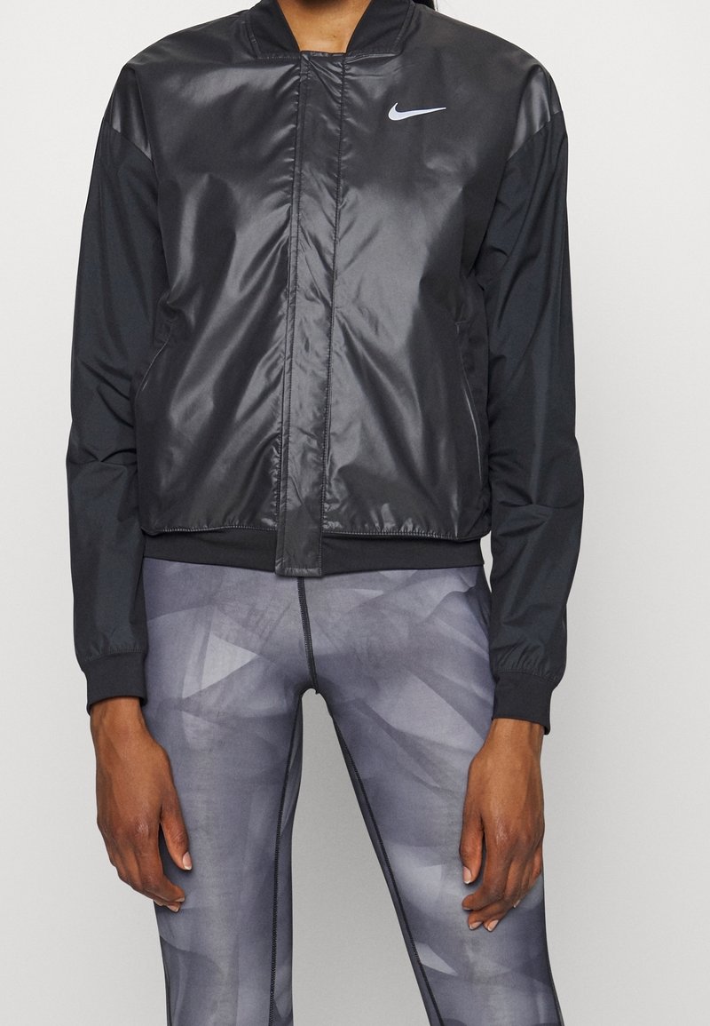 Personne portant une veste légère noire Nike et des leggings gris à motif géométrique, debout devant un fond clair uni.