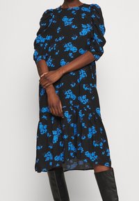 Černé květinové šaty s modrými květinovými vzory, nabíranými rukávy a vrstvenou sukní. Vyrobené z lehké tkaniny, s volným střihem a zaobleným výstřihem.