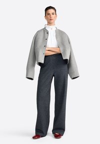 Giacca grigia corta con grandi bottoni, camicia bianca con volant, pantaloni larghi grigio scuro e scarpe bordeaux.