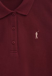 Bordeauxrode poloshirt met een gestructureerde gebreide stof, drie knopen en een klein geborduurd figuurlogo in licht beige op de borst.