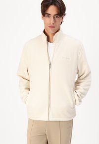 Cream fleece jacket with high collar, front zipper, two side pockets, and embroidered logo. Soft texture, relaxed fit, suitable for layering.