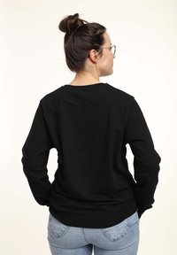Mujer con cabello oscuro recogido en un moño, que lleva gafas, una camisa negra de manga larga y jeans azul claro, vista desde atrás sobre un fondo liso.