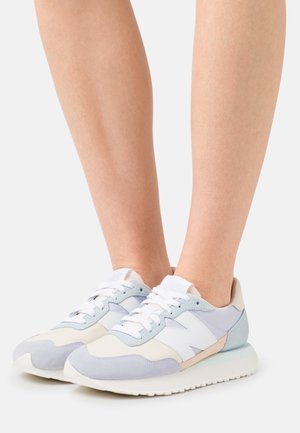 Personne portant des baskets New Balance de couleur pastel avec des lacets blancs, debout sur une surface blanche.