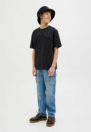 Camiseta de algodón negra con logo rojo, combinada con jeans cargo azul claro y zapatos de cordones marrón oscuro. Un gorro tipo bucket negro completa el look.