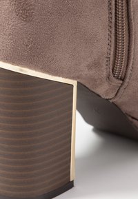 Botín de ante marrón con tacón rectangular adornado en dorado y detalle de cremallera. El tacón tiene una textura acanalada y una base oscura.