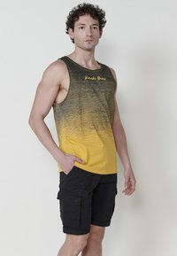 Rumena in črna gradient majica brez rokavov z logotipom, v kombinaciji s črnimi cargo kratkimi hlačami z stranskimi žepi in zavihanimi robovi, oboje iz tkanine.