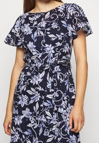 Kvinna klädd i en marinblå klänning med vitt och ljuslila blommönster och korta fladderärmar, knuten i midjan med ett matchande bälte.