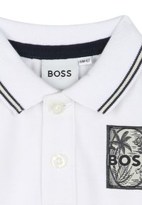 White polo shirt with a contrasting navy collar featuring two stripes. The shirt has a badge with palm tree graphics and "BOSS" text.