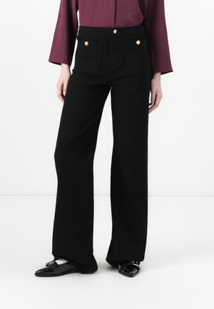 Femme portant un pantalon noir à jambes larges avec poches plaquées devant et boutons dorés, assorti à des chaussures noires pointues et un haut bordeaux.