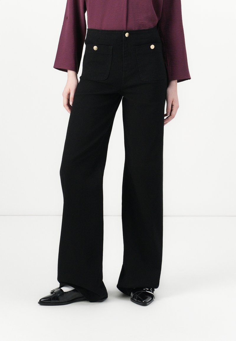 Femme portant un pantalon noir à jambes larges avec poches plaquées devant et boutons dorés, assorti à des chaussures noires pointues et un haut bordeaux.