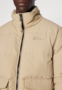 Beige pufferjack met hoge kraag, ritsluiting en voorkruiszakken. Kenmerkt een gestructureerde stof en het Jack Wolfskin-logo op de borst.