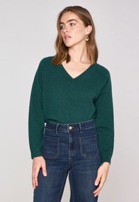 Maglione verde a v di lana morbida con maniche lunghe, abbinato a jeans blu scuro a vita alta con chiusura a bottone e tasche posteriori.