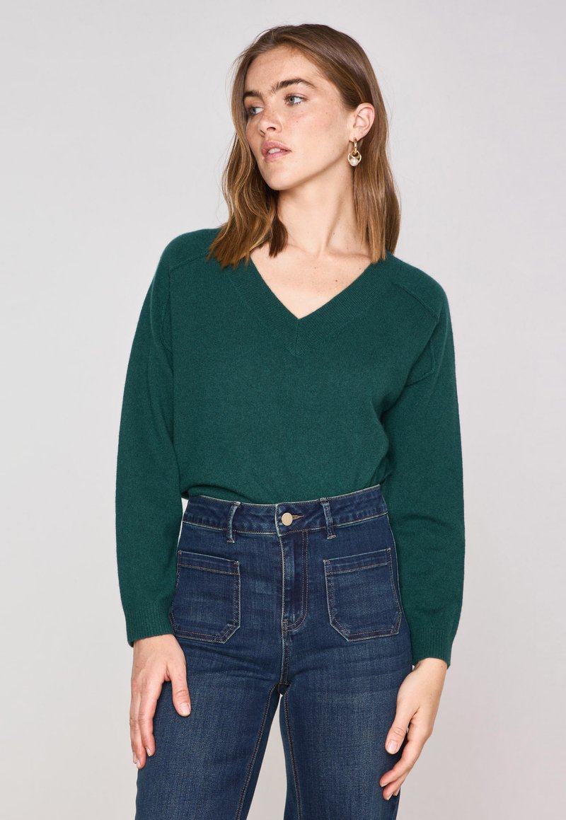 Maglione verde a v di lana morbida con maniche lunghe, abbinato a jeans blu scuro a vita alta con chiusura a bottone e tasche posteriori.