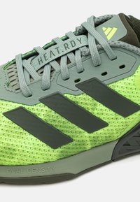 Scarpa sportiva grigia e verde con tomaia in rete, tre strisce nere, etichetta "HEAT.RDY" e dettagli testurizzati. Presenta una suola a doppia densità.