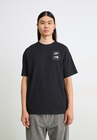 T-shirt en coton noir à manches courtes et à col rond. Présente un petit logo blanc sur la poitrine. Porté avec un pantalon à rayures grises.