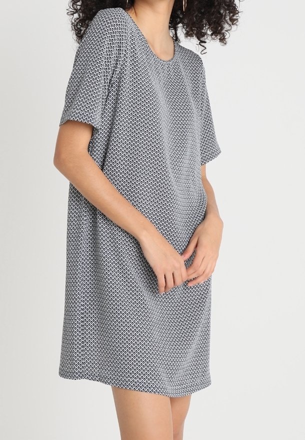 Femme aux cheveux bouclés portant une robe ample à manches courtes à motif noir et blanc, debout devant un fond uni.