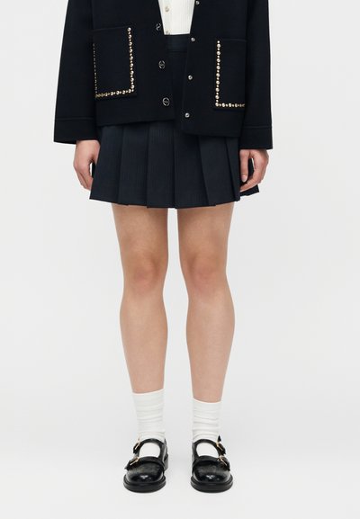 Άτομο που φοράει μαύρο τζάκετ με τρουκς, μαύρη πλισέ φούστα, λευκές κάλτσες και μαύρα παπούτσια Mary Jane με αγκράφα, στέκεται μπροστά σε απλό φόντο.