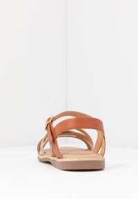 Sandalia de cuero marrón con múltiples correas y una hebilla dorada, presenta una suela plana con un brillo texturizado dorado a lo largo del borde.