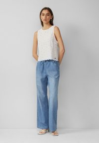 Bijela bezrukavna majica s uzorkom rupica i valovitim rubom, uparen sa svijetloplavim širokim trapericama i bijelim sandalama na petu.