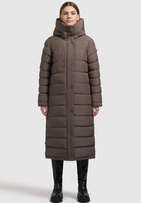 Lång, puffig brun kappa med hög krage, framfickor och knappstängning. Materialet verkar vara slätt med en kviltad textur.