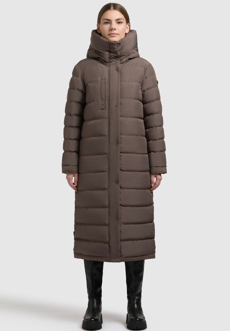 Lång, puffig brun kappa med hög krage, framfickor och knappstängning. Materialet verkar vara slätt med en kviltad textur.