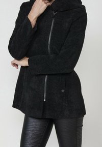 Personne portant une veste zippée noire texturée avec une capuche, les mains placées près du visage et de la taille, associée à un pantalon en cuir synthétique noir.